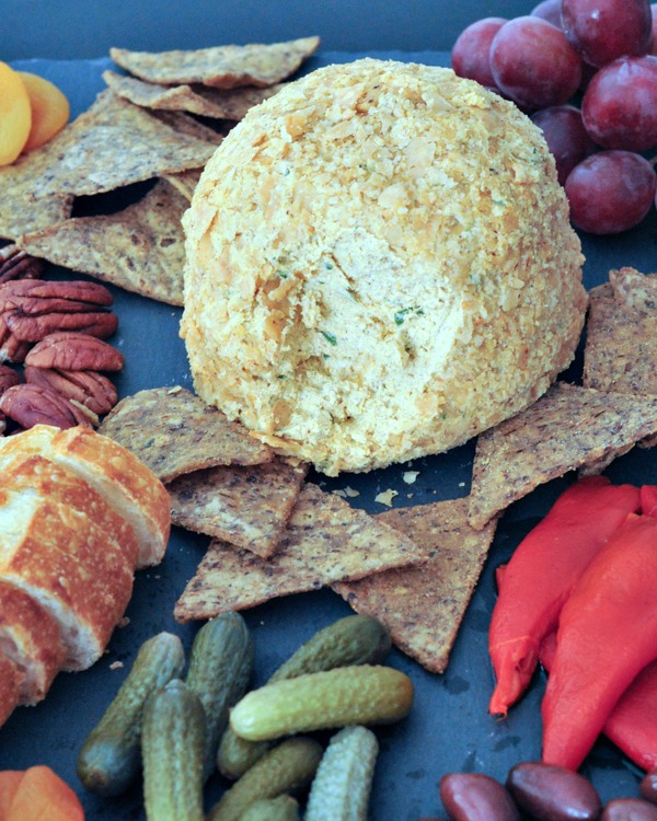 close up of tan colored Spiced Pumpkin Basil Cheese Ball on a black slate board with crackers, pecans, capers, sliced baguette, purple grapes, cornichon pickles,and olives