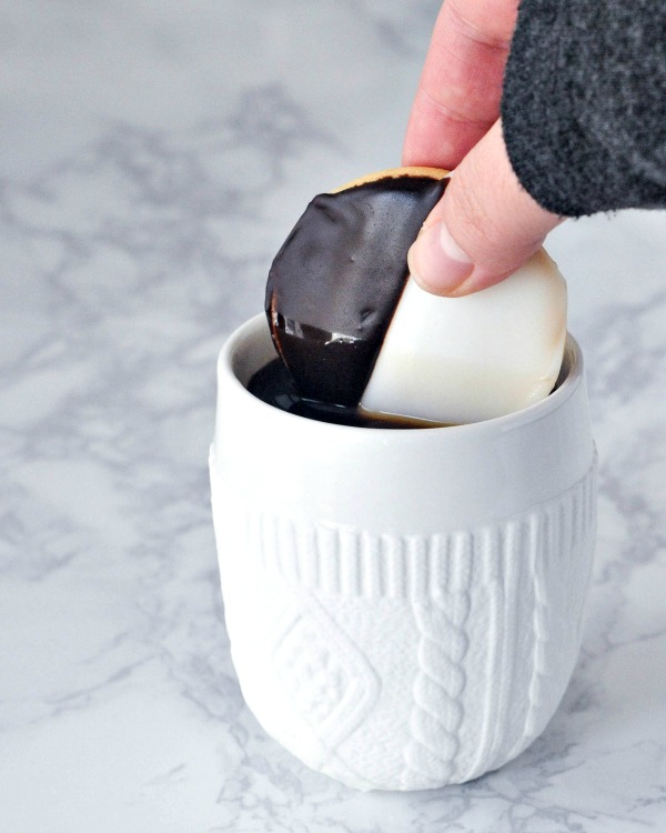 a Black and White Cookie being dunked into a white mug of coffee. mug is white matte ceramic with a cable knit sweater pattern. a black and white cookie is a cake like vanilla cookie with half chocolate frosting and half white frosting.