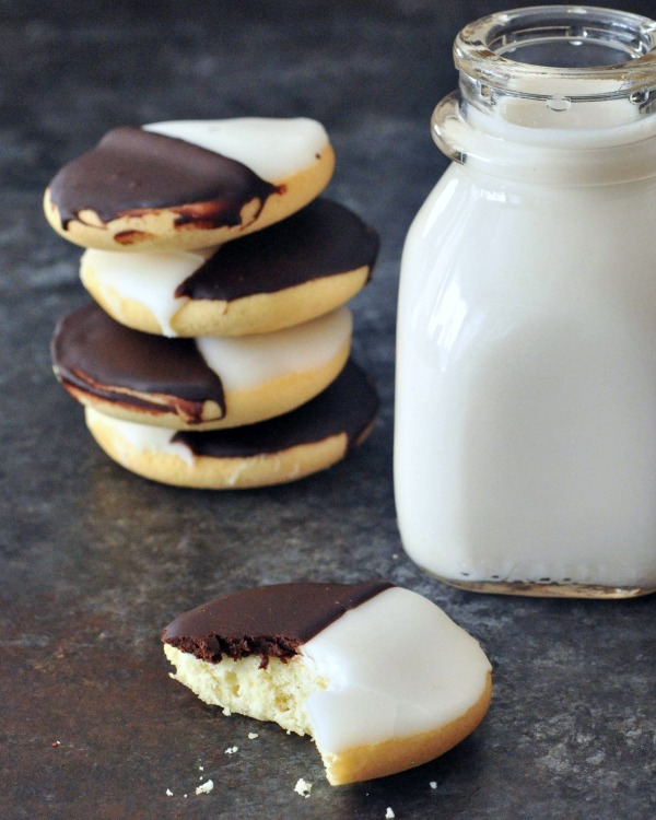 Black and White Cookies stacked four high, next to a single cookie with a bite out of it and a short glass bottle of milk. black and white cookie is a cake like vanilla cookie with half chocolate frosting and half white frosting.