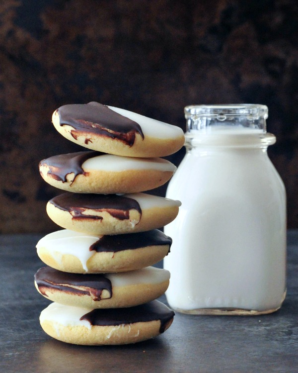 Black and White Cookies stacked six high, next to a short glass bottle of milk. black and white cookie is a cake like vanilla cookie with half chocolate frosting and half white frosting.