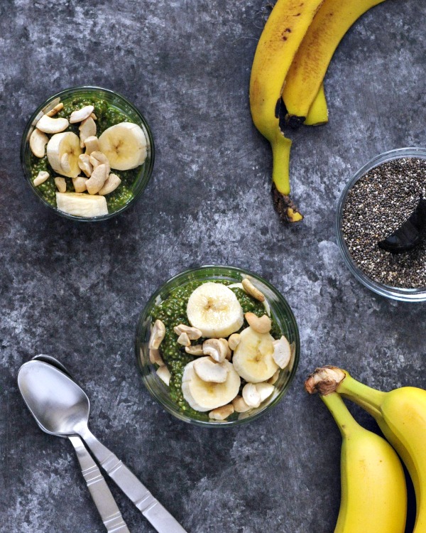 Green from added spinach, pineapple chia pudding in a glass with banana slices. unpeeled bananas and a bowl of dry chia seeds on the side.