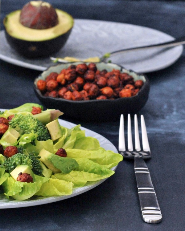 A salad of fresh greens, broccoli, and avocado slices is garnished with smoky sweet chickpeas. A bowl of extra chickpeas sits next to the salad with a half avocado.