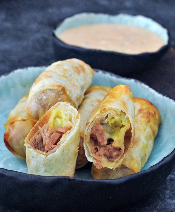 reuben rolls (jackfruit reuben sandwich filling wrapped in egg roll wrapper) in a shallow blue bowl on a darker blue background, smaller blue bowl of thousand island dressing on the side.