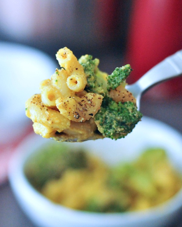 a spoon holding one bite of broccoli mac and cheese, with the bowl of mac and cheese blurred in the background.