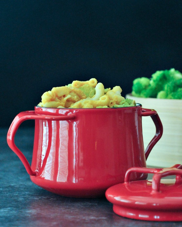 One serving of broccoli mac and cheese in a red serving bowl, with a bamboo steamer basket full of broccoli.