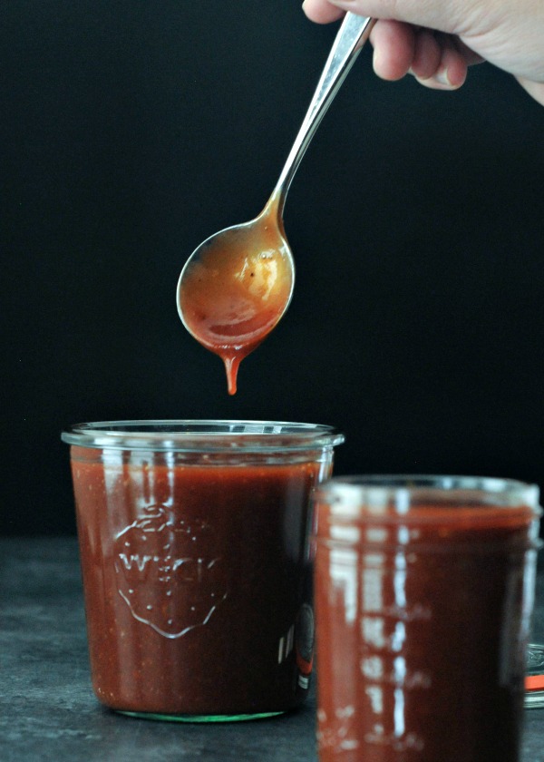 two glass jars of a gluten free vegan BBQ sauce sitting on a dark grey marble countertop, with a hand holding a spoon coated in BBQ sauce.