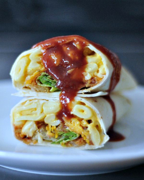 A BBQ mac and cheese burrito sliced in half on a white plate, with BBQ sauce dripping on top.