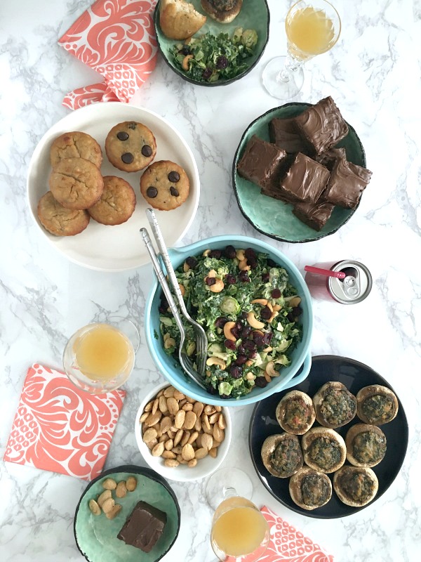 Overhead view of a brunch table: a bowl of lemon brussels and kale salad with cashews and dried cranberries, a plate of muffins, a plate of mini quiche, a bowl of nuts, and glasses of champagne