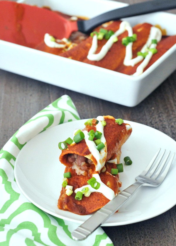 one Bean and Cauliflower Rice Enchilada stacked on a second one, on a white plate next to a green and white cloth napkin, and a full pan of red sauce enchiladas in the background.