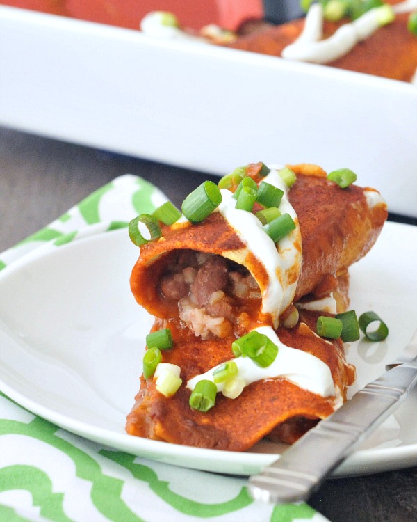 one Bean and Cauliflower Rice Enchilada stacked on a second one, on a white plate next to a green and white cloth napkin, and a full pan of red sauce enchiladas in the background.