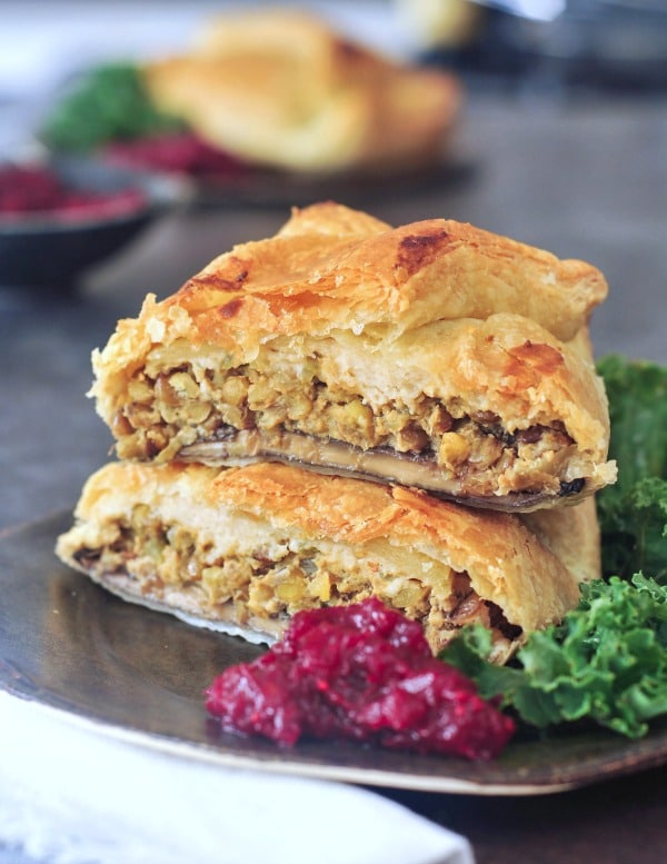 Individual portobello wellington cut in half and stacked on a dark grey plate to show inside filling of lentil veggie stuffing, kale salad and bright red cranberry sauce on the side
