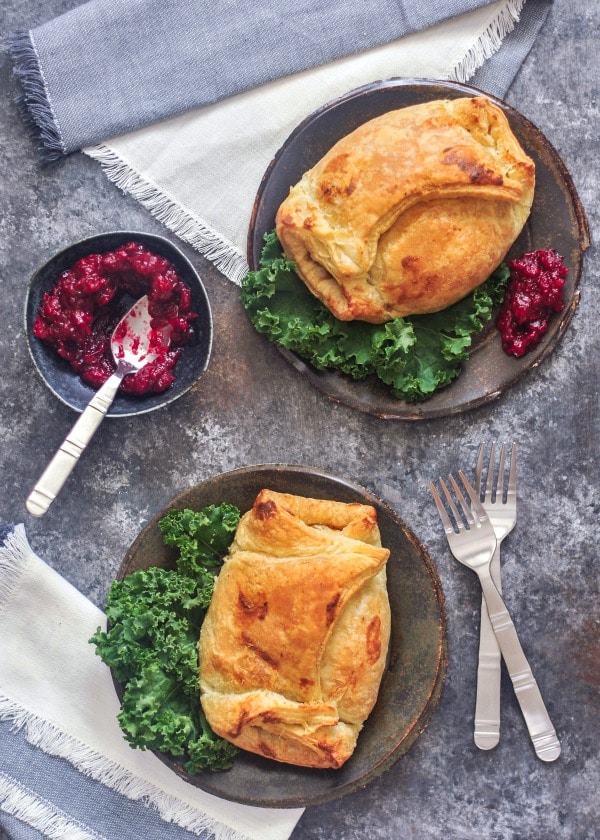 overhead view of two individual portobello wellingtons on dark grey plates, kale salad and bright red cranberry sauce on the side