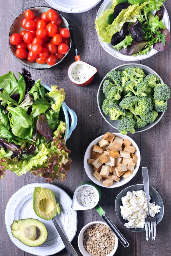 overhead view of a build your own salad bar ingredients in bowls: broccoli, tomatoes, leafy greens, tofu cubes, avocado slices, nuts, vegan cheese, and dressing.