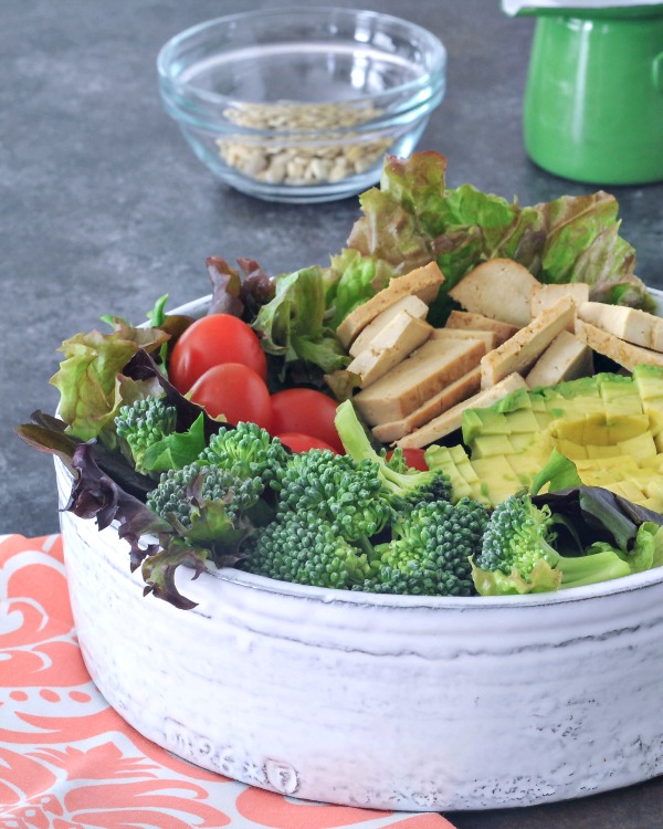 a large serving bowl of salad: greens, broccoli, tomatoes, tofu slices, avocado, nuts in a bowl on the side.