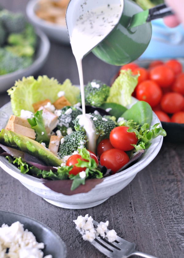 ranch dressing being poured over a salad in a bowl: greens, tofu cubes, tomatoes, broccoli