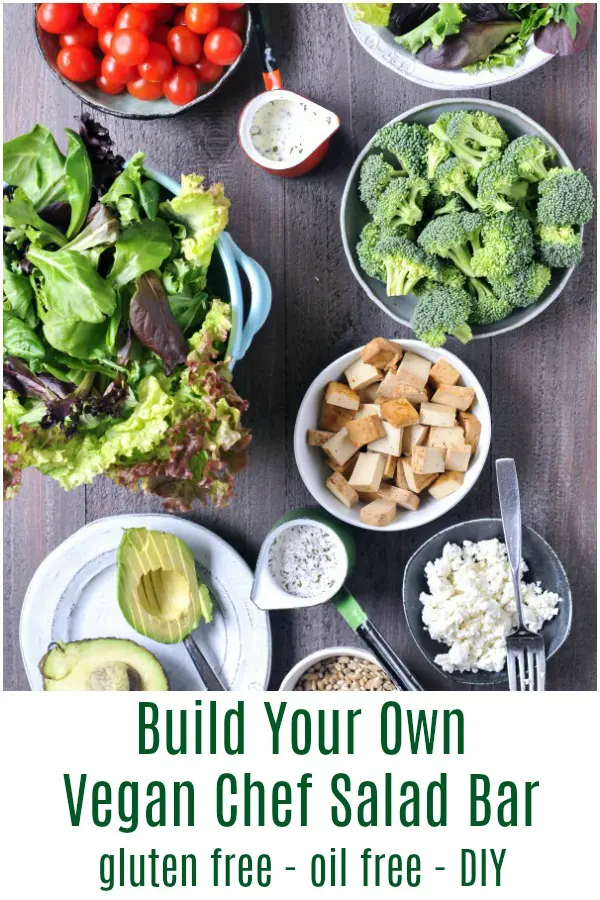 overhead view of a build your own salad bar ingredients in bowls: broccoli, tomatoes, leafy greens, tofu cubes, avocado slices, nuts, vegan cheese, and dressing.