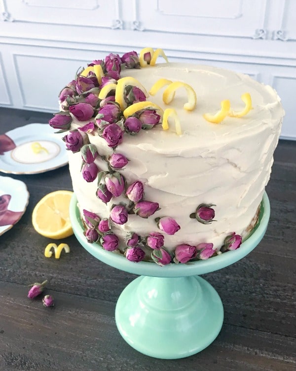 overhead view of full Lemon Elderflower Layer Cake on a light green cake stand; cake garnished with pink edible dried flowers and yellow curled lemon peel