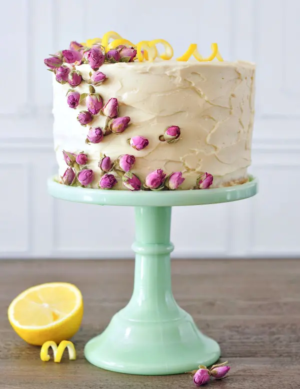 Lemon Elderflower Layer Cake on a light green cake stand, cake garnished with pink edible dried flowers and yellow lemon peel curls