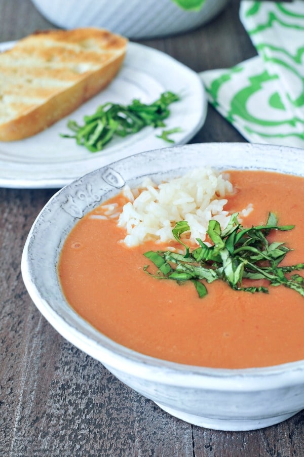 Tomato orange soup in a rustic white bowl, garnished with chopped basil and white rice, a plate with crusty baguette on the side.