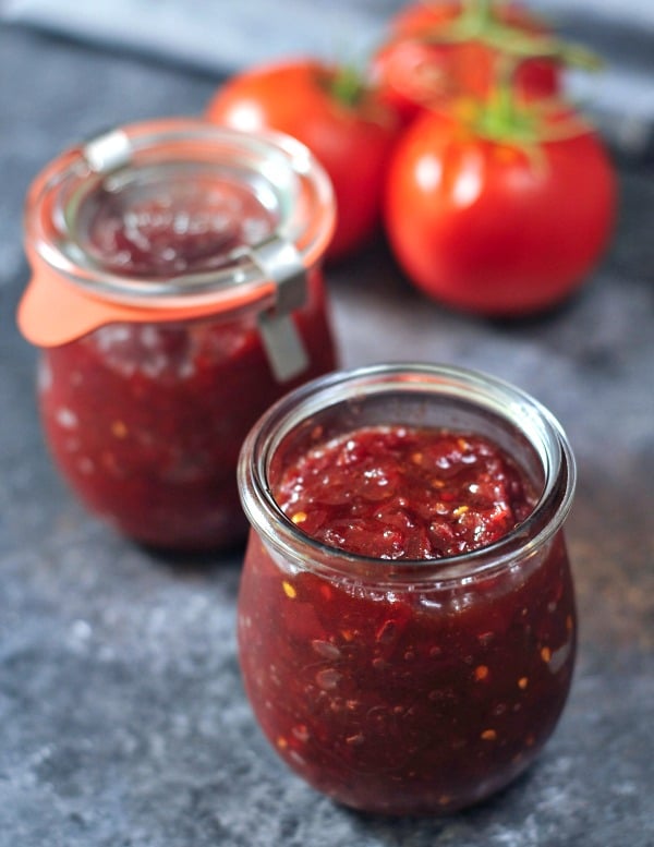 two small glass jars of tomato jam on dark grey marble surface, fresh tomatoes in background