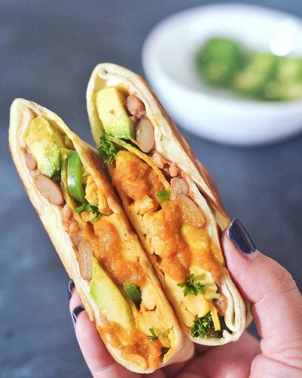 One ranchero style breakfast crunchwrap sliced in half and held in a hand against a dark blue background. Crunchwrap is filled with scrambled egg, beans, ranchero sauce, jalapenos.