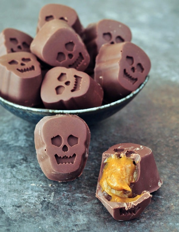 a bowl full of bite size, skull shaped Halloween chocolates stuffed with pumpkin caramel. one outside of the bowl is cut open to show the golden caramel inside.