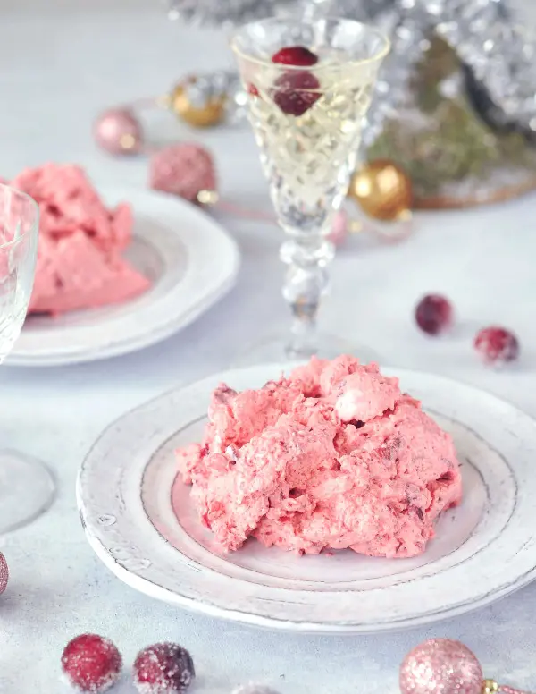 single servings of bright pink cranberry fluff jello salad on rustic white plates, sugared cranberries and champagne glasses in the background