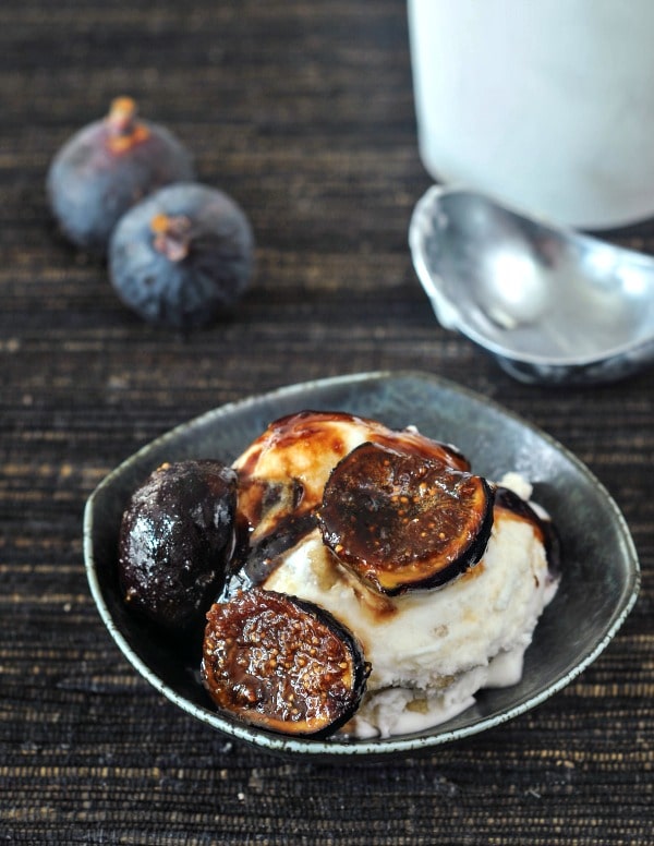 Espresso sugared figs in a rustic grey bowl over ice cream, two whole figs, a carton of ice cream, and an ice cream scoop in background.