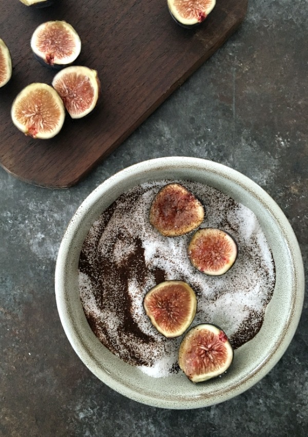 Sliced figs in a shallow bowl with espresso powder and sugar, with a cutting board and more sliced figs on the side