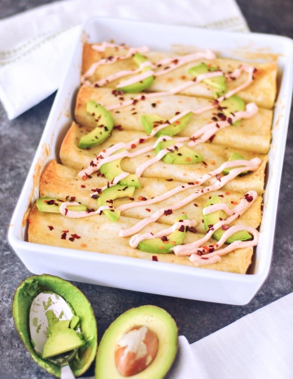 Buffalo Jackfruit Enchiladas in a white baking dish, topped with avocado slices, a drizzle of sour cream, and red chile flakes. a half avocado and an empty shell of avocado with a spoon sits next to enchiladas.