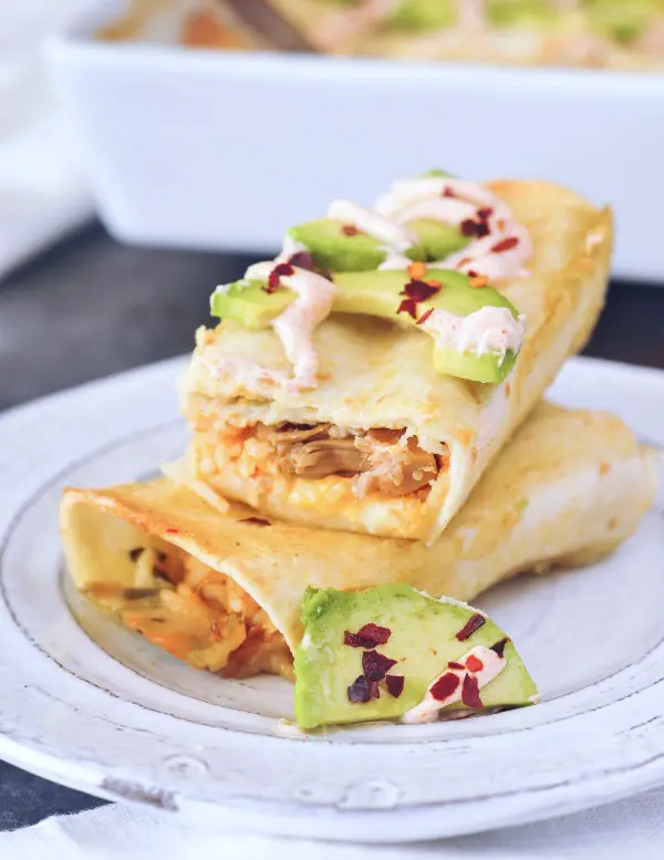 Vegan Buffalo Jackfruit Enchiladas sliced in half and stacked on a grey rustic plate, insides filled with orange buffalo sauced, shredded jackfruit, rice, topped with avocado slices and red chile flakes.