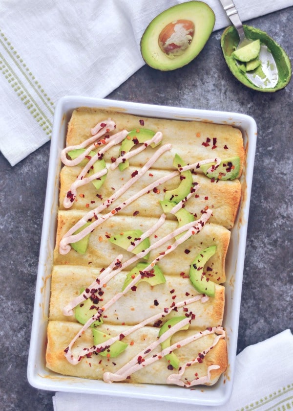 overhead view of Buffalo Jackfruit Enchiladas in a white baking dish, topped with avocado slices, a drizzle of sour cream, and red chile flakes