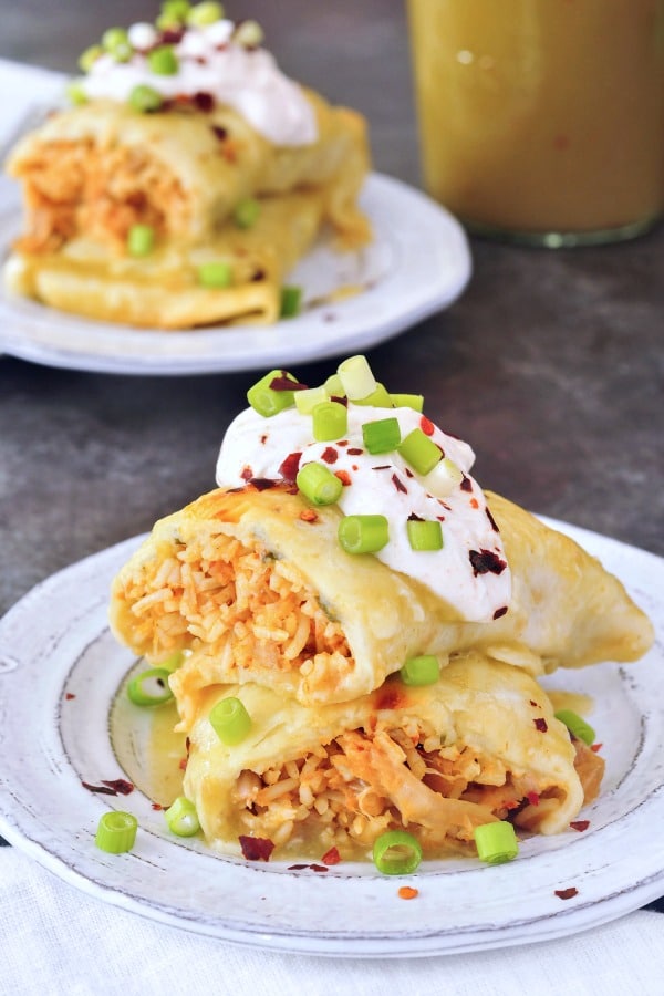 Vegan Buffalo Jackfruit Enchiladas sliced in half and stacked on a grey rustic plate, insides filled with orange buffalo sauced, shredded jackfruit, rice, topped with sour cream and sliced green onion.