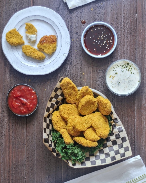 Vegan chicken nuggets in a shallow bowl with black and white checkered paper and kale garnish. dipping sauces on the side (ranch, teriyaki sauce, tomato)