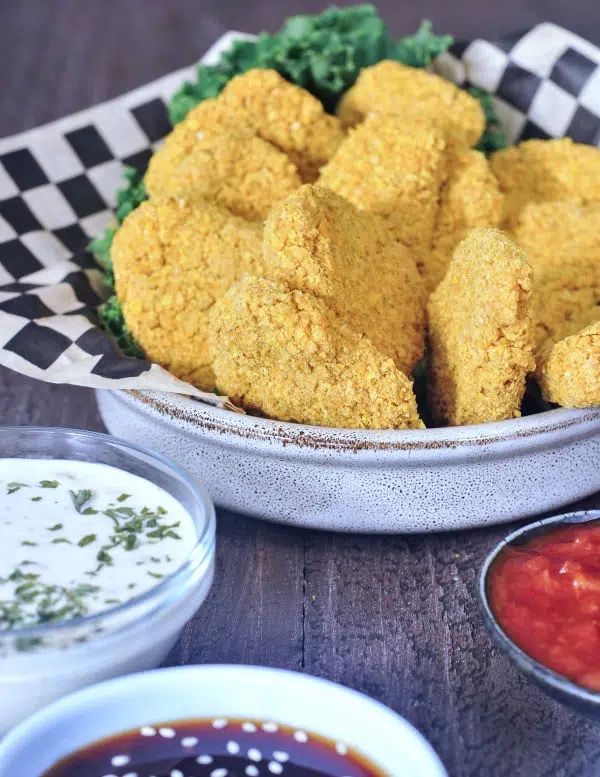 Vegan chicken nuggets in a shallow bowl with black and white checkered paper and kale garnish. dipping sauces on the side (ranch, teriyaki sauce, tomato)