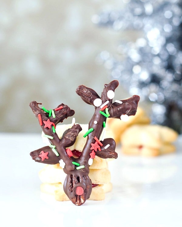 a chocolate covered pecan and pretzel reindeer propped up against a stack of shortbread cookies, silver holiday tree in background