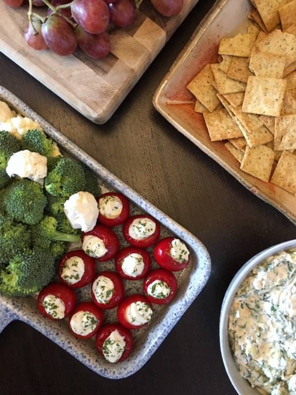 overhead view of round red peppadew sweet peppers filled with herb cheese (Boursin style) on a blue and white speckled tray next to a wood board with purple grapes, a wooden square dish with crackers, and a bowl of spinach dip.
