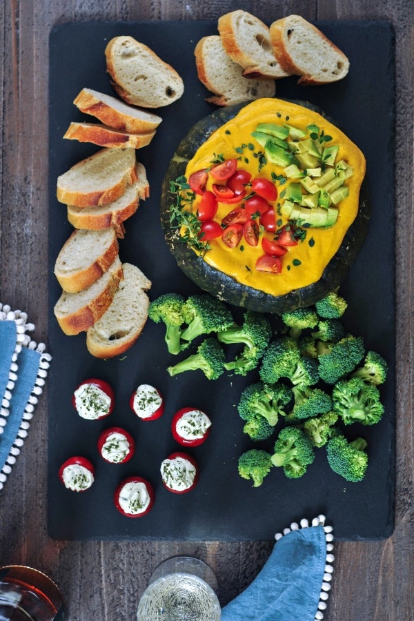 overhead view of bright yellow Spicy Chipotle Kabocha Dip served in the dark green squash shell, topped with chopped avocado and tomatoes and minced herbs. sliced baguette, broccoli trees, and stuffed tomatoes served alongside.