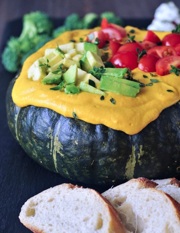 bright yellow Spicy Chipotle Kabocha Dip served in the dark green squash shell, topped with chopped avocado and tomatoes and minced herbs. sliced baguette bread served alongside.