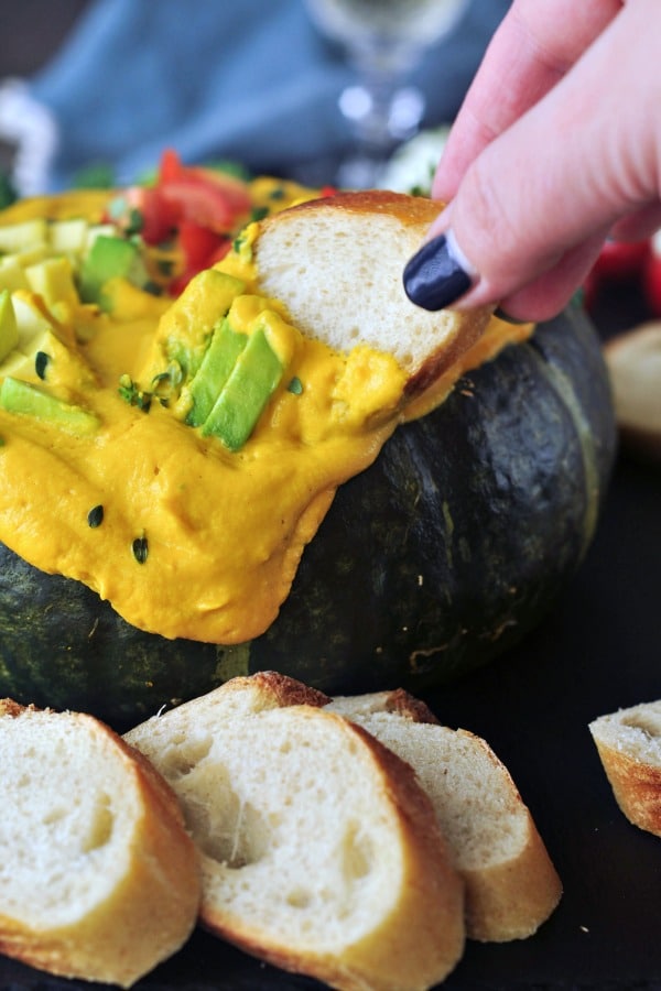 a hand dipping a baguette slice into bright yellow Spicy Chipotle Kabocha Dip, which is served in the dark green squash shell, topped with chopped avocado and tomatoes and minced herbs. sliced baguette bread served alongside.