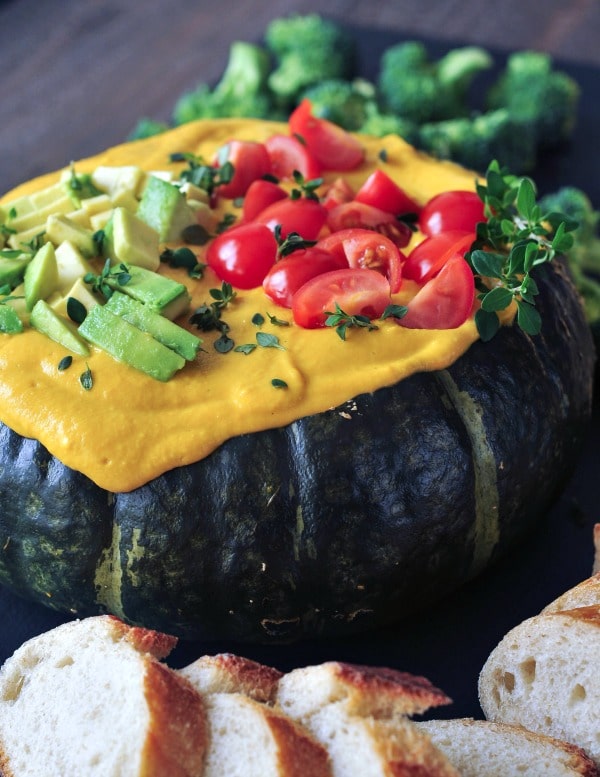 bright yellow Spicy Chipotle Kabocha Dip served in the dark green squash shell, topped with chopped avocado and tomatoes and minced herbs. sliced baguette bread served alongside.