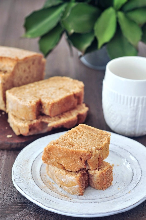 one slice of snickerdoodle bread sliced in half and stacked, the remaining full loaf in background next to a white ceramic mug.