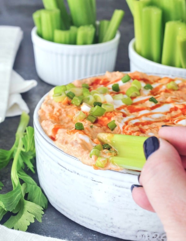 a hand dipping a piece of celery into a vegan buffalo chicken dip in a large straight sided, rustic grey serving bowl, garnished with hot sauce and sour cream drizzle and sliced green onion. set on a dark grey marble tabletop, with smaller bowls of celery slices for dipping.