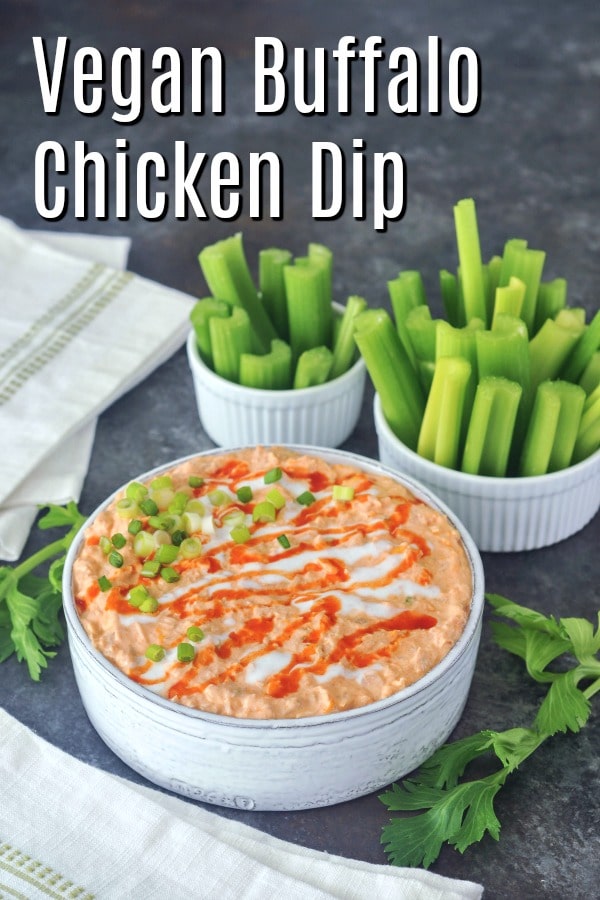 Vegan buffalo chicken dip in a large straight sided, rustic grey serving bowl, garnished with hot sauce and sour cream drizzle and sliced green onion. set on a dark grey marble tabletop, with smaller bowls of celery slices for dipping.