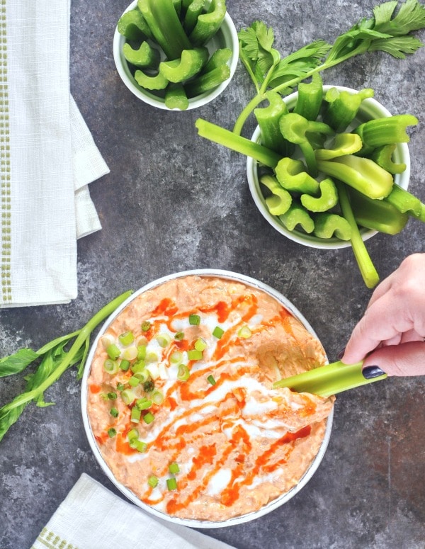 overhead view of vegan buffalo chicken dip in a large straight sided, rustic grey serving bowl, garnished with hot sauce and sour cream drizzle and sliced green onion. set on a dark grey marble tabletop, with smaller bowls of celery slices for dipping.
