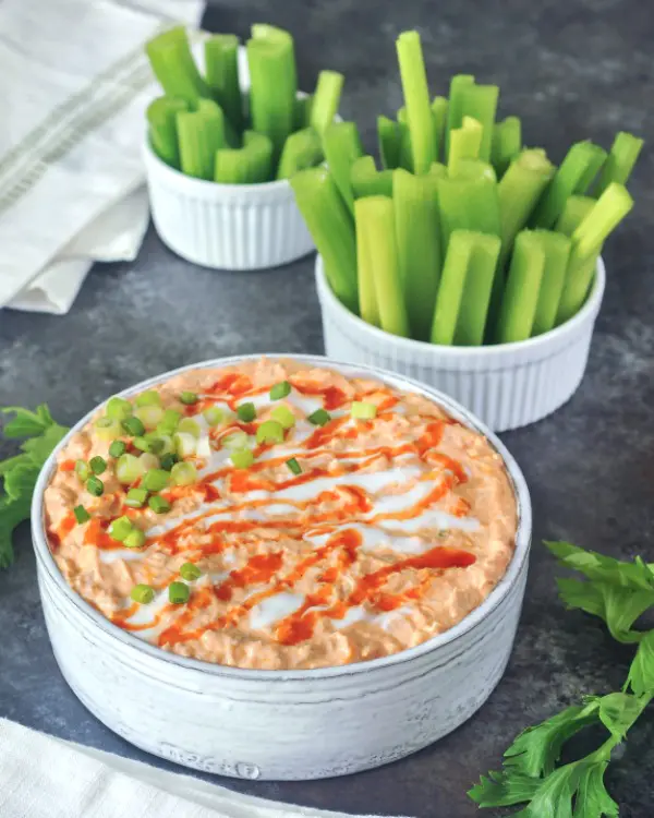 Vegan buffalo chicken dip in a large straight sided, rustic grey serving bowl, garnished with hot sauce and sour cream drizzle and sliced green onion. set on a dark grey marble tabletop, with smaller bowls of celery slices for dipping.