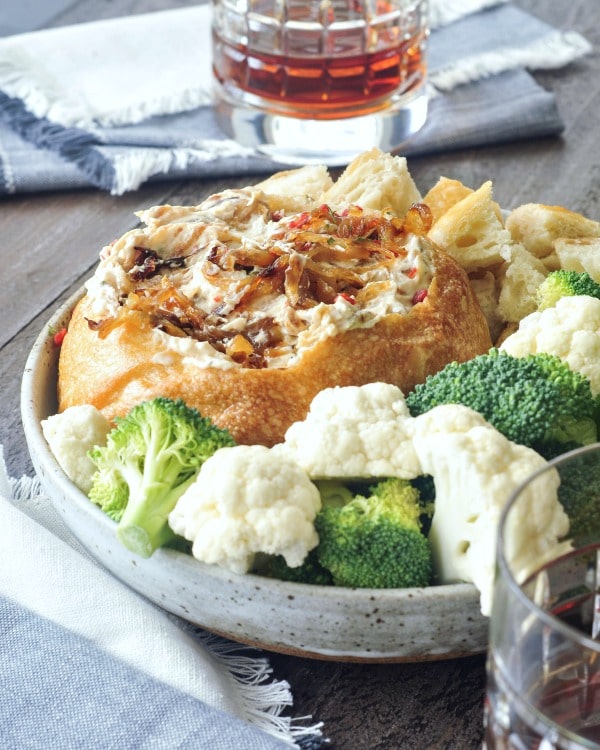 Warm Caramelized Onion Bacon Dip served in a rustic bread bowl in a shallow bowl, surrounded by pieces of bread, cauliflower and broccoli trees, celery pieces