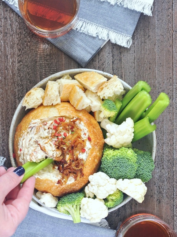 overhead view of Warm Caramelized Onion Bacon Dip served in a rustic bread bowl in a shallow bowl, surrounded by pieces of bread, cauliflower and broccoli trees, celery pieces