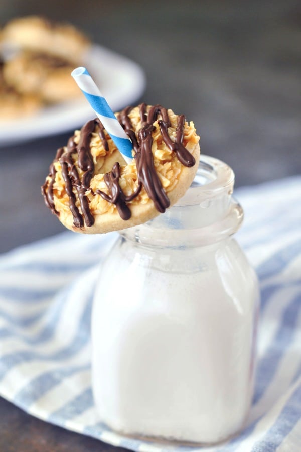 a single Samoa Girl Scout Cookie (shortbread with coconut caramel and a chocolate drizzle) sitting on top of a small glass milk bottle, with a blue and white striped straw through the hole of the cookie