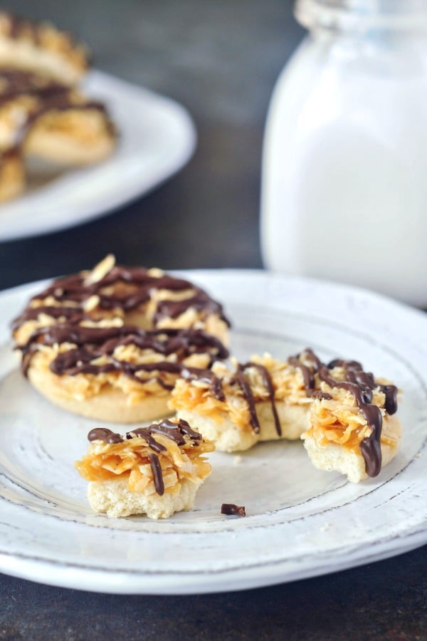 two Samoa Girl Scout Cookies (shortbread with coconut caramel and a chocolate drizzle) on a white rustic plate, with a small glass bottle of milk in background.